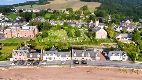 Drone footage steadily advances over a coastal Scottish village, highlighting a stone church, cemetery, and surrounding homes in bright natural daylight