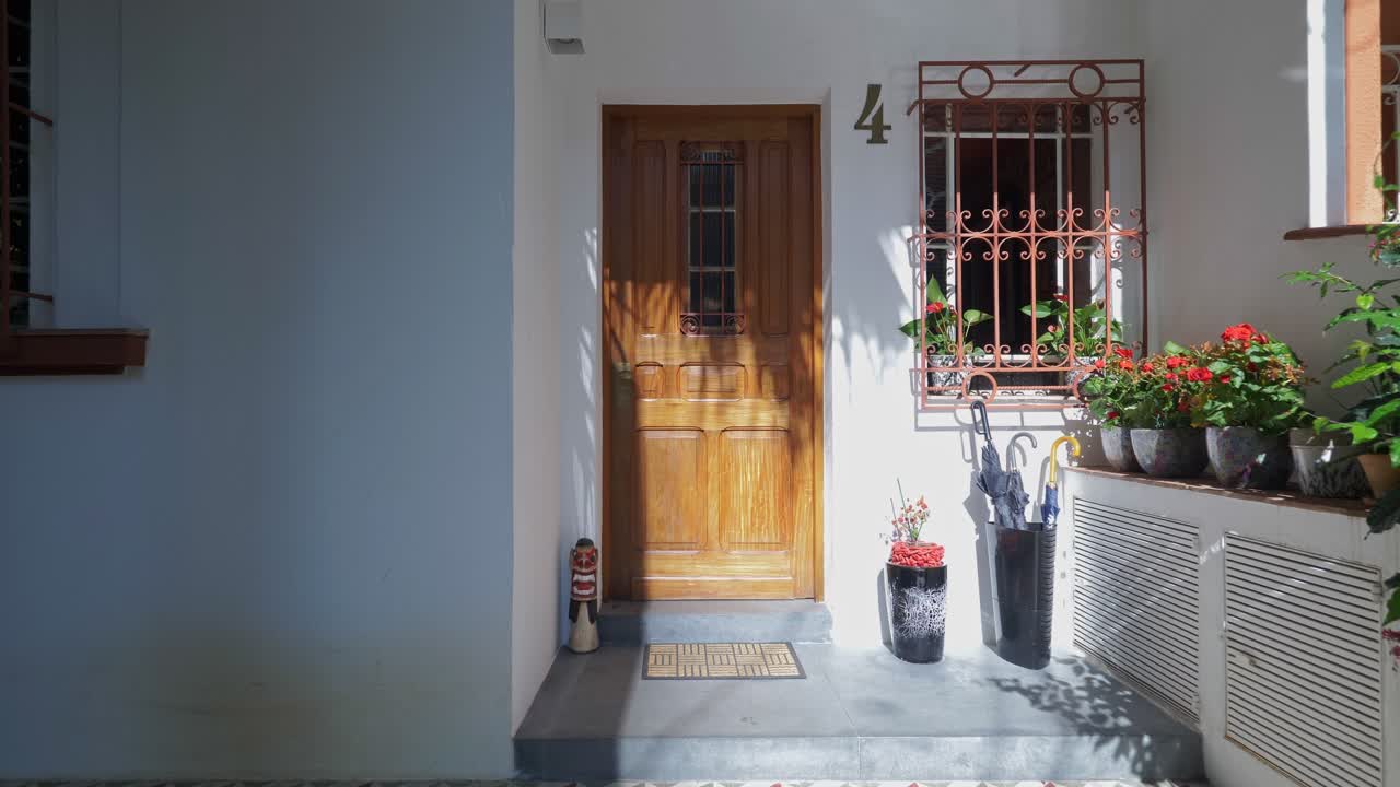A wooden door is partly open on a white wall, next to a flower-filled planter, umbrellas, and a decorative window grille, creating a bright and cheerful entryway.