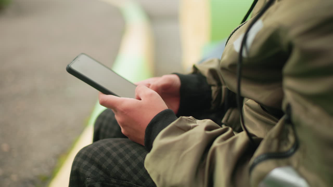 Close up rear view of kid in jacket holding smartphone with both hands, sitting outdoors deeply engaged in device operation, showing focus and concentration on digital screen