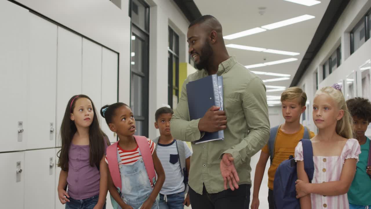 Video of happy african american male teacher and diverse pupils walking at school hall