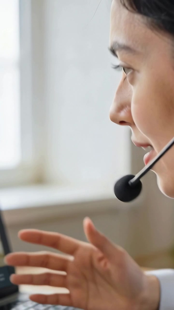 Video verticale: Donna che parla al telefono, in top, seduta alla scrivania, usa cuffie e laptop per lavoro.