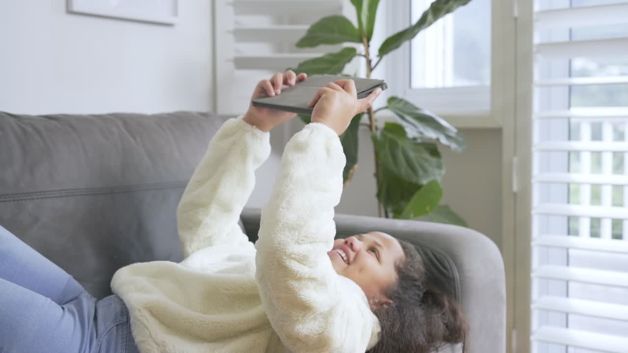 Child girl tapping and swiping tablet on grey sofa under shuttered window, laughing after wobble