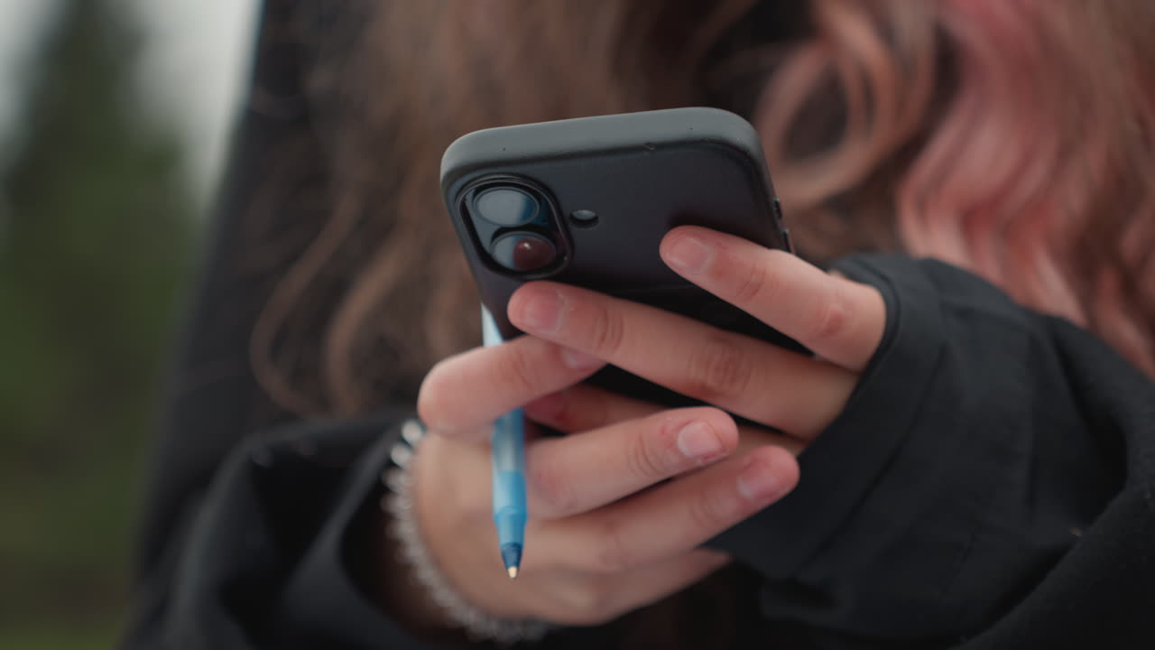 Primo piano di una studentessa che tiene in mano telefono e penna, mani femminili con capelli rosa e braccialetto di perle che indossa una felpa nera con cappuccio, sfondo verde sfocato all'aperto, controllo dei messaggi mentre prepara gli appunti, calma e concentrata