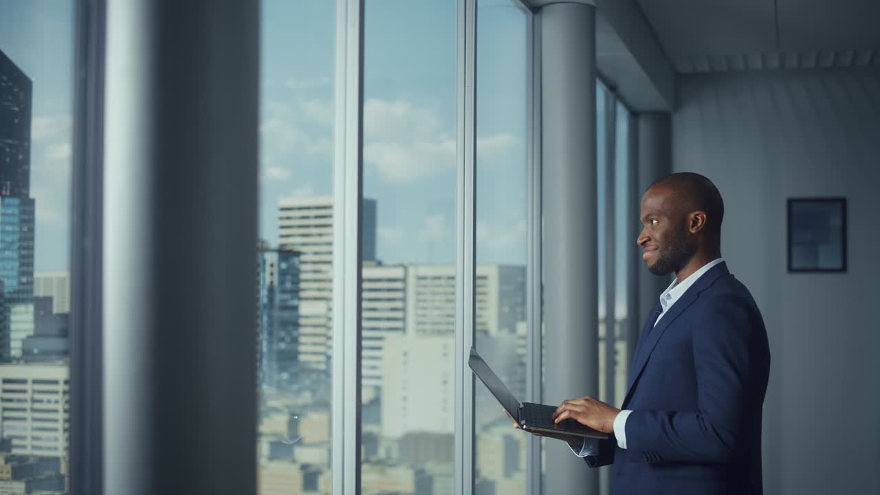 Thoughtful Black Businessman in a Tailored Suit Using Laptop while Standing in Office Near Window on Big City. Successful Corporate Top Manager Doing Data Analysis for e-Commerce Startup