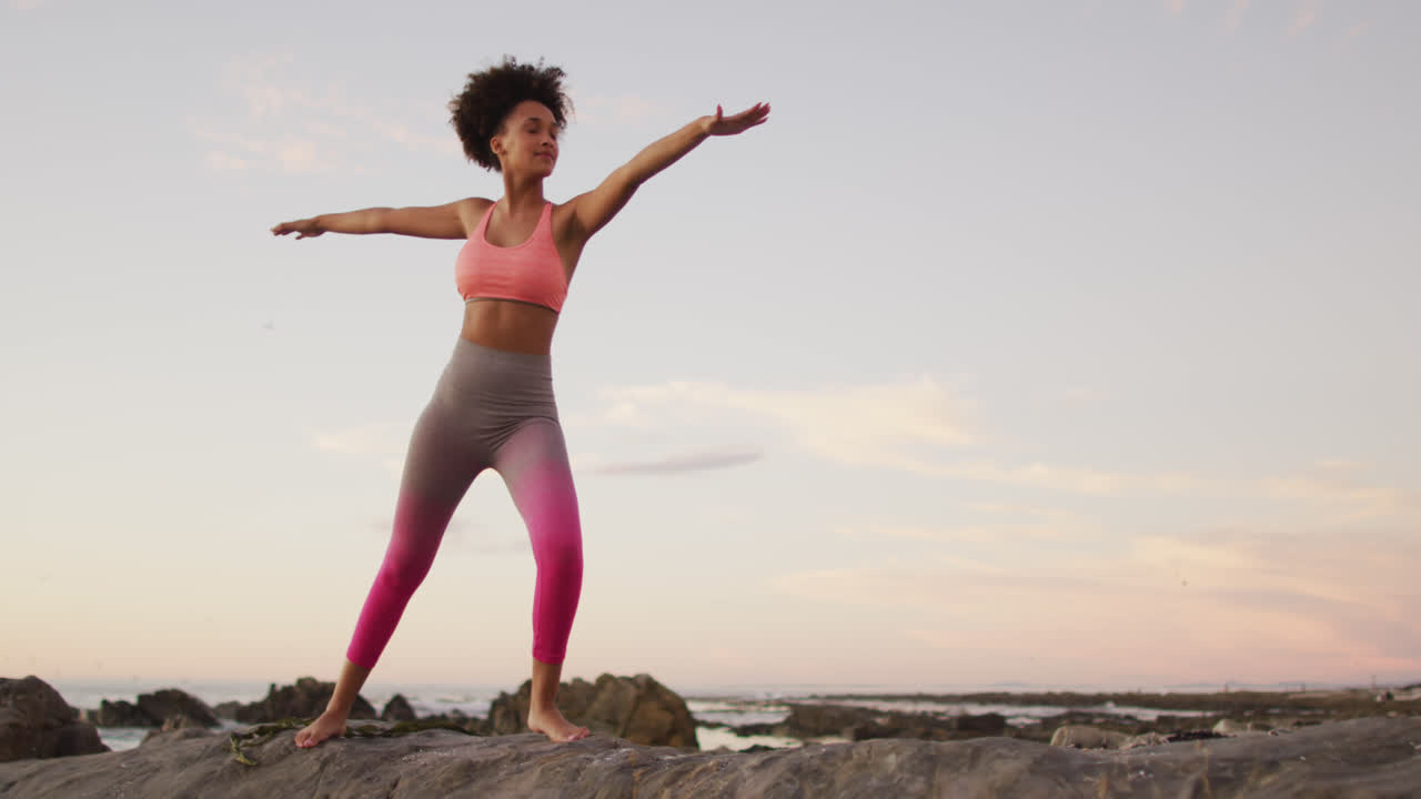 African american woman practicing yoga and meditating on the rocks near the sea during sunset