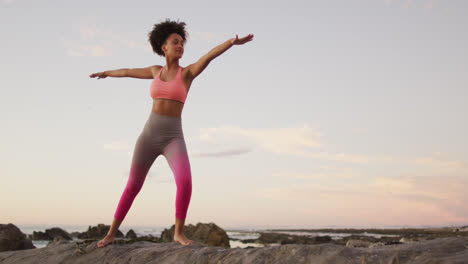 African-american-woman-practicing-yoga-and-meditating-on-the-rocks-near-the-sea-during-sunset