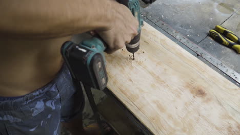 a shirtless worker drilling hole into a wooden board while making a skateboard