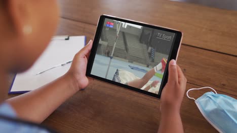 compuesto de una mujer sentada en una mesa con una máscara facial, viendo el evento de salto alto de atletismo en la tableta