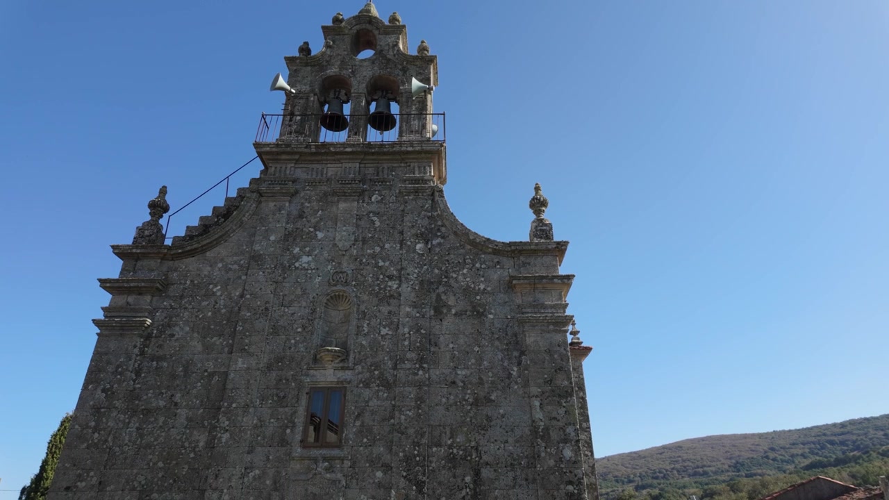 Vista dal basso della chiesa di Santa María de Porqueira a Ourense, Galizia, che mostra la sua storica facciata in pietra, un campanile a vela ornato e la tradizionale architettura religiosa sotto un cielo azzurro e limpido.