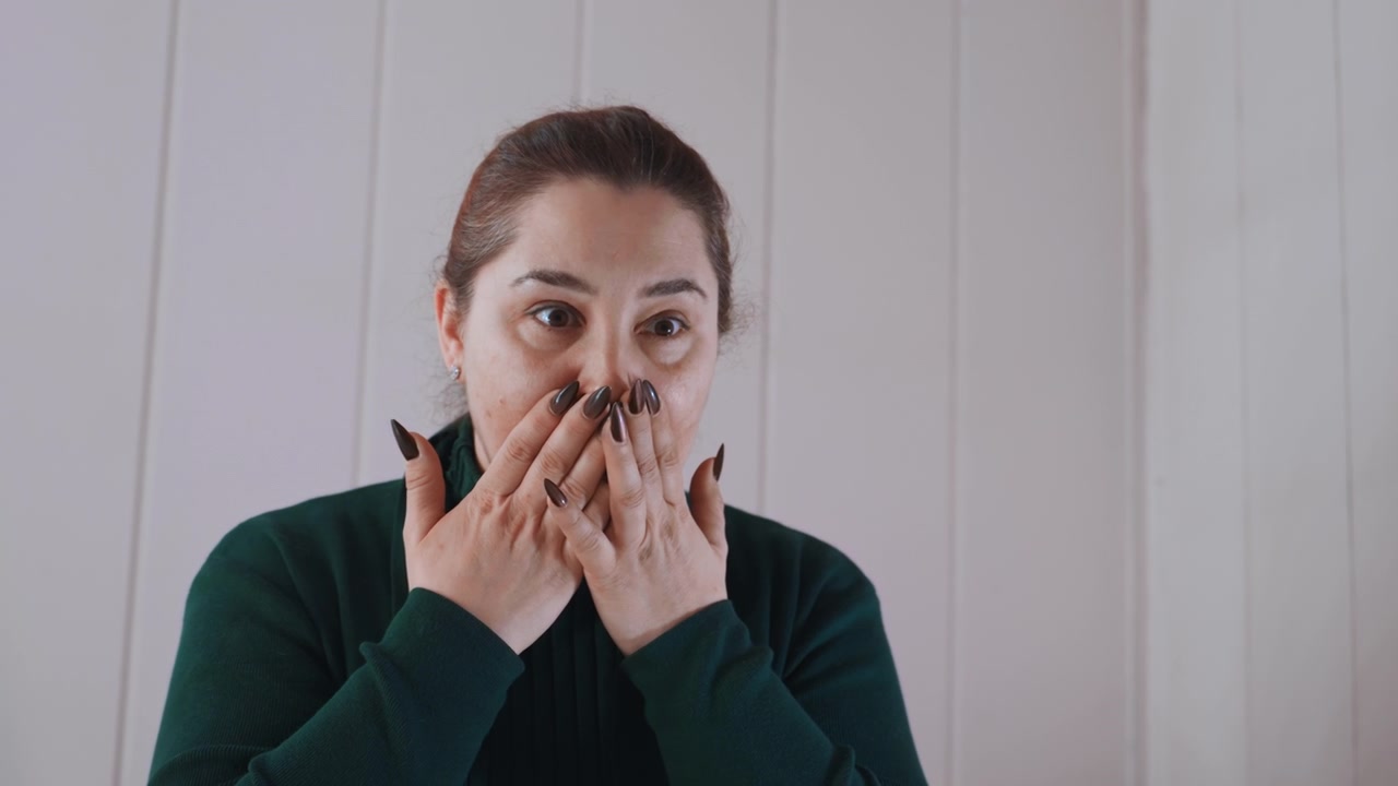 Donna caucasica che si stringe il petto a casa, ansimando e coprendosi la bocca, espressione ansiosa, interni con pannelli, respiro affannoso, che cerca il telefono, disagio emotivo che suggerisce un'emergenza medica.