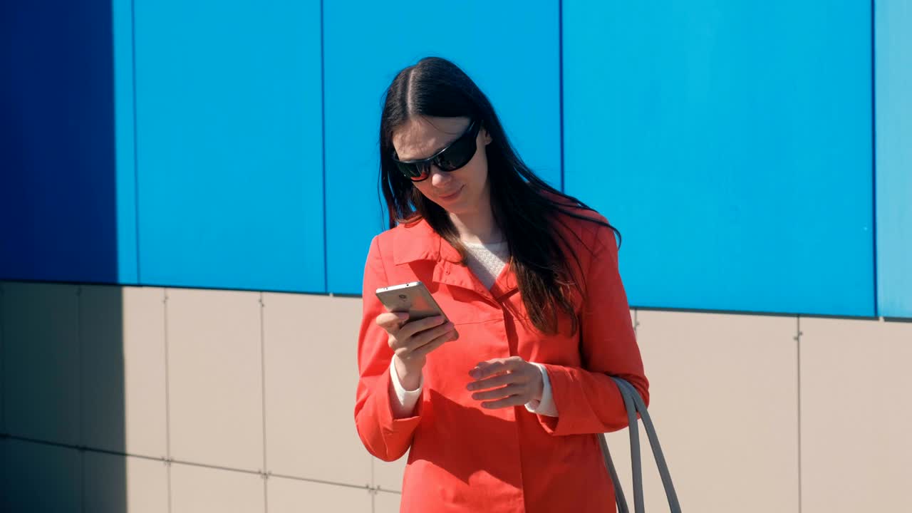 은 코트를 입은 선글라스를 입은 젊은 갈색 여성이 누군가를 기다리고 드폰을 확인하고 문자 메시지를 보내고 있습니다.