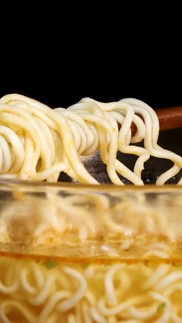 ガラスの器から箸が黄色い麺を持ち上げ、巻き上げている様子を捉えたクローズアップの縦ショット。