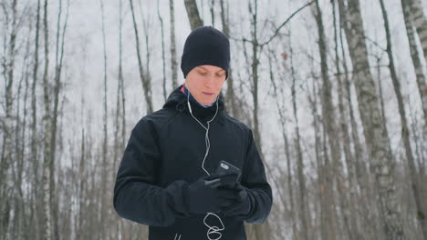 Ein-Junger-Mann-In-Einem-Schwarzen-Hammer-Bereitet-Sich-Im-Winter-Auf-Das-Joggen-Im-Park-Vor,-Enthält-Eine-Laufanwendung-Und-Rennt-Mit-Einem-Sprachassistenten-Durch-Den-Wald,-Um-Geschäfte-Zu-Machen-Und-Einen-Gesunden-Lebensstil-Zu-Führen.