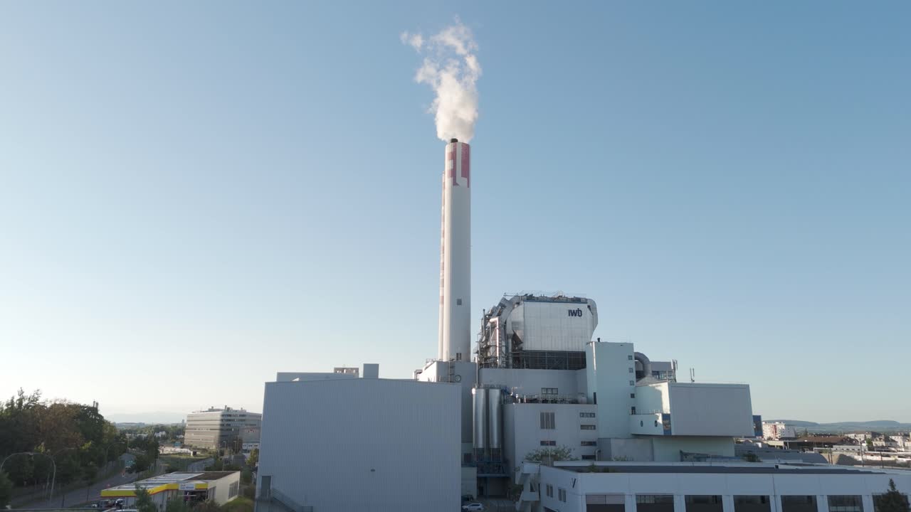 Filmato di un grande stabilimento industriale con un'alta ciminiera che emette fumo, sullo sfondo di un cielo azzurro e circondato da edifici industriali