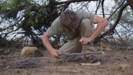 caucasian male survivalist using fire bow and fire drill at camp in wilderness