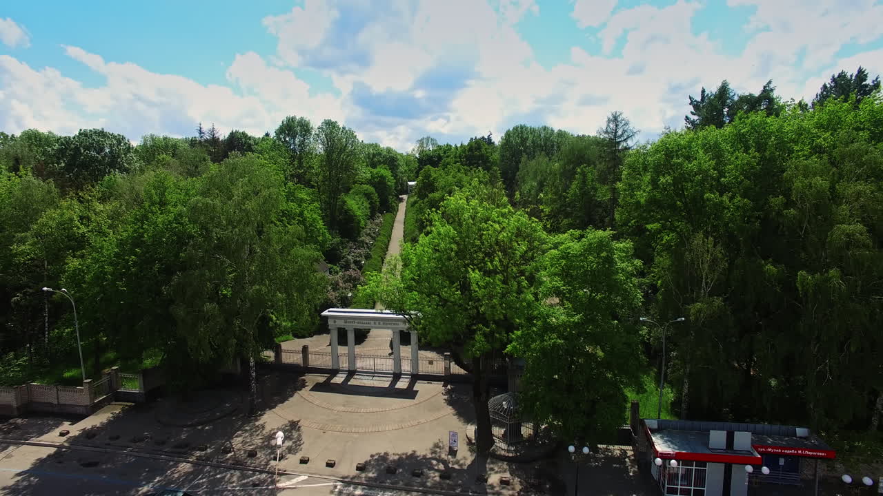 Flying closer to the city park with white columns at the entrance. Long path through the green trees leading to the building. Top view.