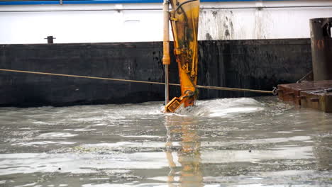 Shot-of-the-shovel-of-an-excavator-on-a-pontoon-next-to-a-ship,-deepening-a-canal-SLOMO