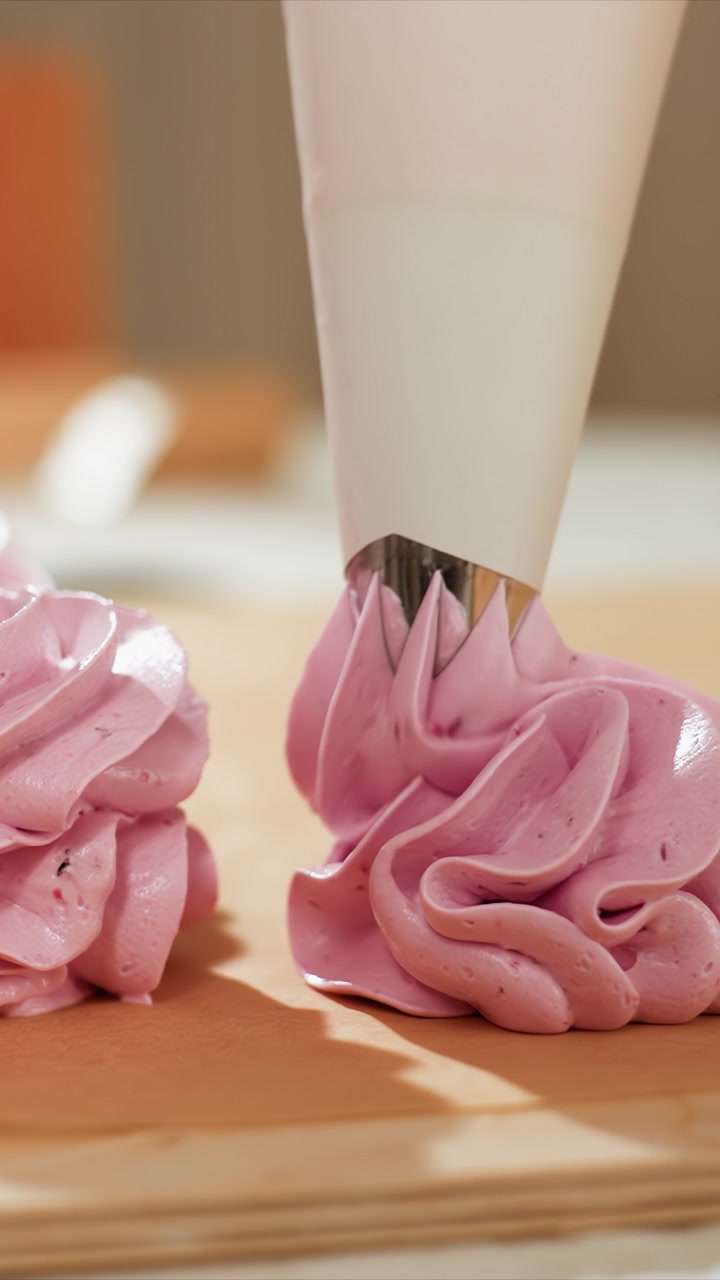 Primo piano di glassa rosa che viene spremuta da una sac à poche bianca su. Texture lucida e consistenza liscia. Lo sfondo in legno suggerisce un ambiente da cucina con strumenti da forno con glassa