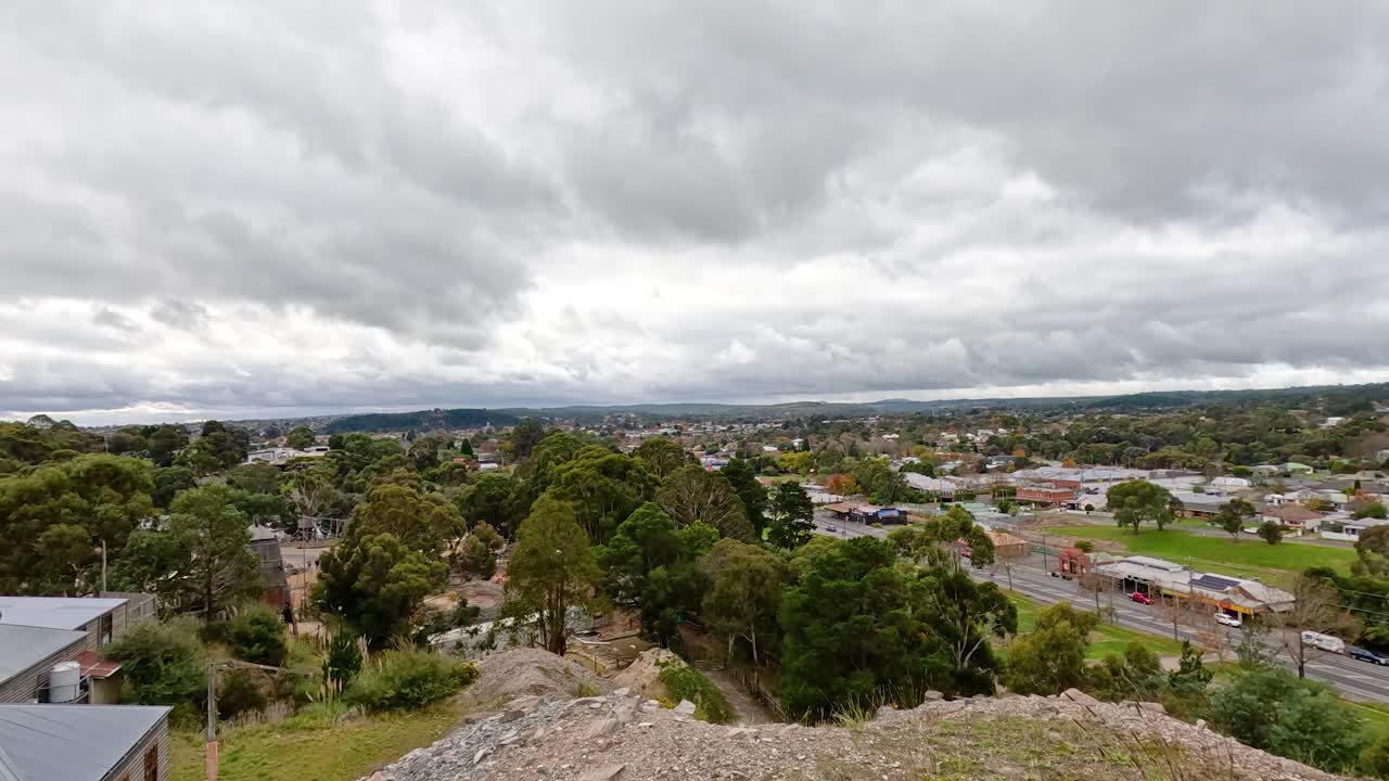 구름이 가득한 하늘 아래 푸른 초록색과 도시 구조물을 보여주는 발라라트의 광활한 공중 풍경, 역동적인 움직임으로 포착되었습니다.