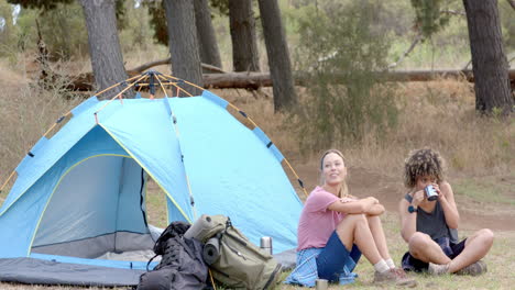 young caucasian woman and biracial woman relax beside a blue tent in the woods