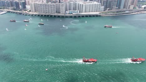 Convoy-of-local-Fishing-boats-causing-in-Hong-Kong-Victoria-bay,-with-city-skyline-in-the-horizon,-Aerial-view