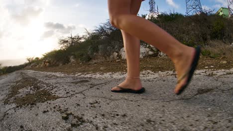 young caucasian female feet tracking shot low to ground over eroded old broken asphalt road, downhill sunset in backrgound