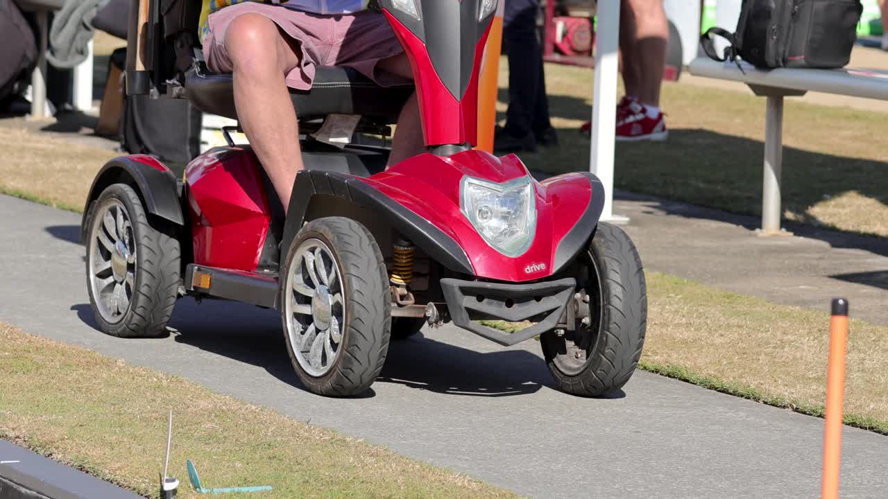 A person drives a red motorized scooter along a sunny outdoor path, surrounded by people and grass