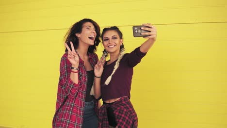 Two-hipster-girls-making-selfie-and-showing-peace-sign-standing-by-the-yellow-wall.-Beautiful-girls-posing-and-taking-photos