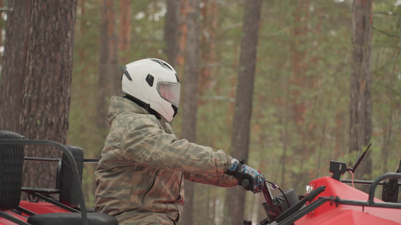 Pilota di quad nella foresta indossa casco bianco e giacca mimetica su un quad rosso, primi piani di profilo che mostrano le mani sull'acceleratore, passo costante lungo i tronchi di pino, luce nuvolosa, atmosfera calma e concentrata.