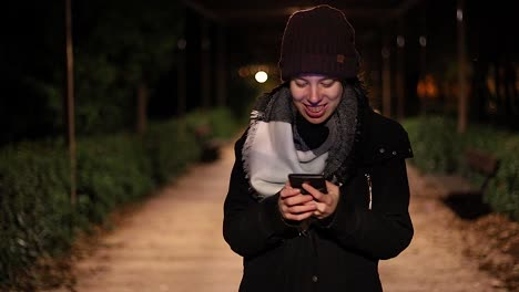 Mujer-Joven-Y-Guapa-Caminando-De-Noche-Y-Mirando-Su-Teléfono-Inteligente-En-Un-Parque