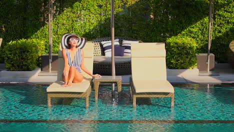 Elegant-woman-with-swimsuit-and-black-and-white-hat-sitting-on-deckchair-in-pool-water