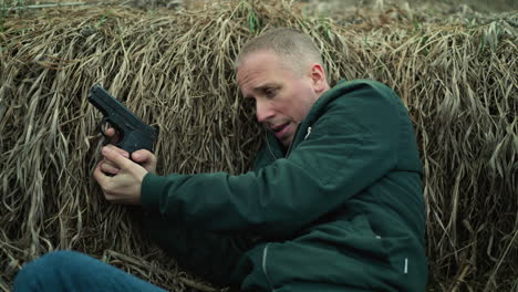 a close view of a man in green jacket and blue jeans holding a pistol, looking and tense while lying against dry grass