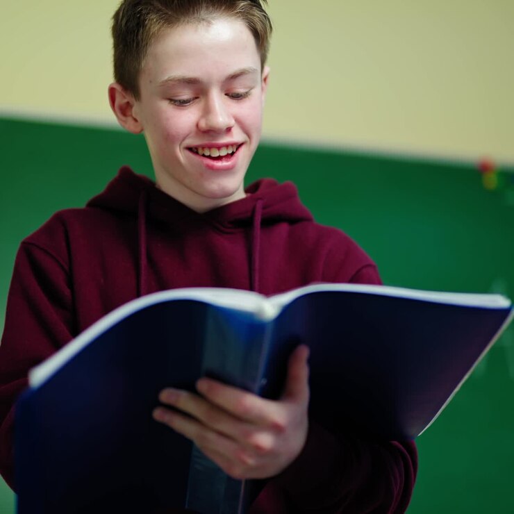 Smiling schoolboy with a folder. Teenage boy standing in the classroom and looking into the school register. Portrait of a happy teenager in the school.