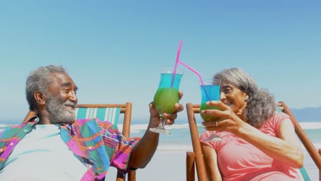 Front-view-of-active-senior-African-American-couple-toasting-drinks-on-deckchair-on-the-beach-4k