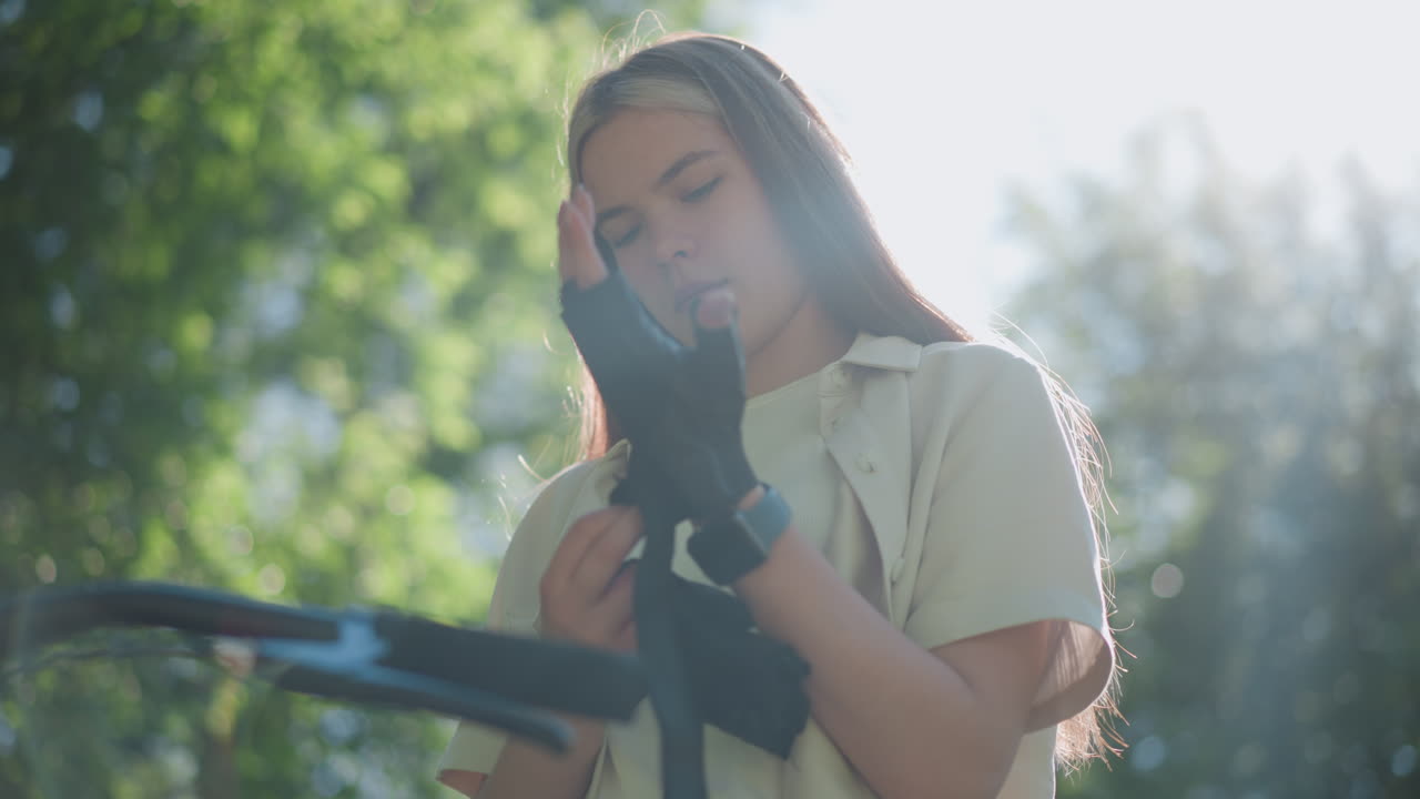 오토바이 장갑을 들고 있는 젊은 여성의 낮은 각도 시각, 왼손에 장갑을 착용할 준비, 집중된 표정, 배경에 흐릿한 녹색 잎자루