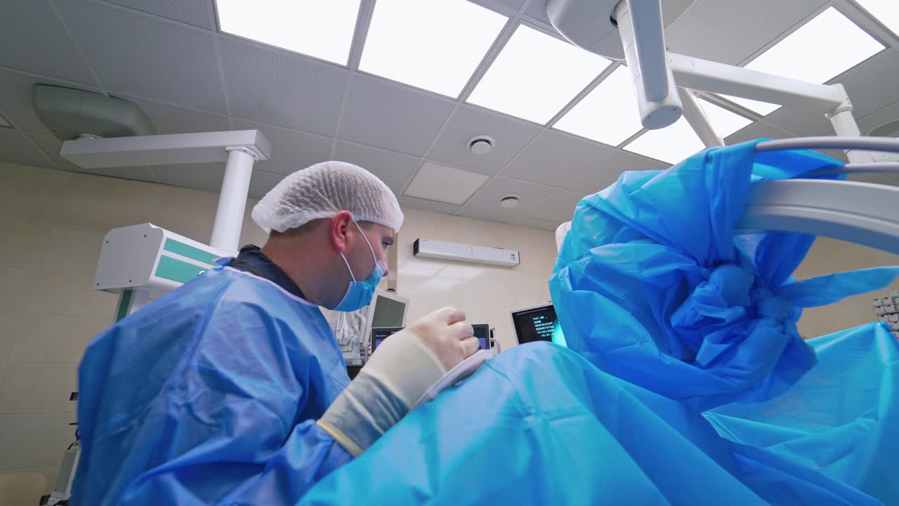 Side view of a surgeon in modern operating room. Professional doctor in medical uniform conducts an operation in private clinic with contemporary equipment.