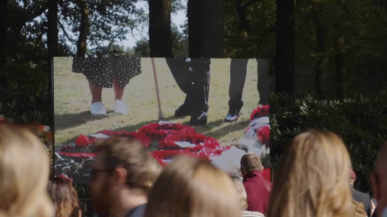 Crowd gathered at Oosterbeek War Cemetery watching a large screen display of live military memorial ceremony, commemorating fallen soldiers. Location: Oosterbeek, Gelderland, Netherlands