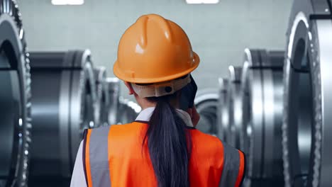 vista de atrás de una ingeniera asiática con casco de seguridad hablando en un teléfono inteligente mientras está de pie en una fábrica de metal