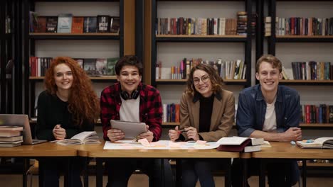 Dos-Jóvenes-Estudiantes-Europeos-Y-Dos-Mujeres-Sentados-A-La-Mesa-Con-Libros-Y-Una-Computadora-Portátil-En-La-Biblioteca-Y-Sonriendo-Alegremente.-Estanterías-En-La-Biblioteca-Con-Muchos-Libros-Al-Fondo.-Camara-Lenta