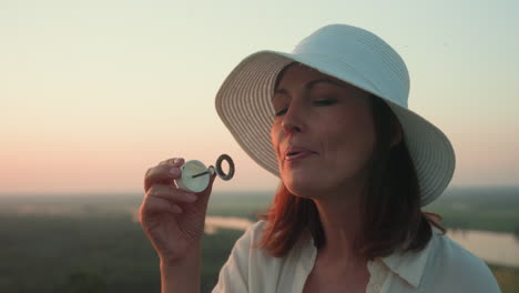 young woman in white wide brim hat blowing delicate soap bubbles into pastel sunset sky over river valley and lush grassy hillside, her profile lit by warm golden light