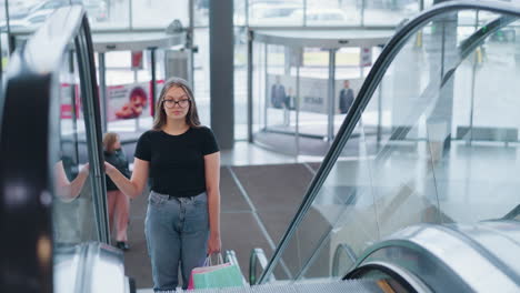 young woman in black top and jeans ascends escalator in mall holding two shopping bags, with other shoppers blurred in the background, reflecting an everyday urban scene