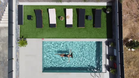 Drone-flies-over-an-outdoor-pool-in-bird's-eye-view---Woman-in-swimming-costume-swims-in-supine-position-on-the-water-surface---Villa-in-Greece-Crete-with-palm-trees