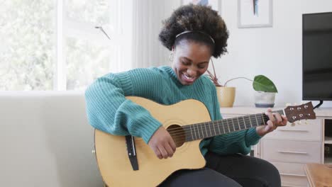 happy african american woman playing guitar and singing sitting on couch at home, slow motion