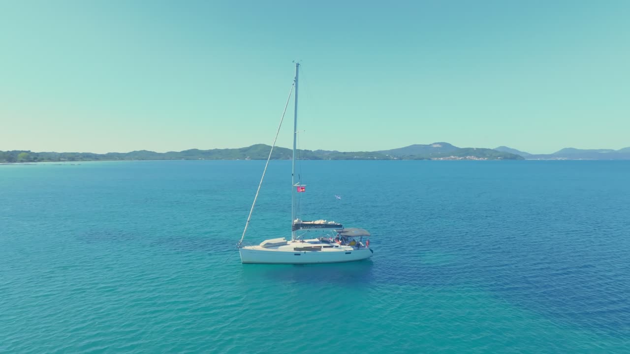 vista panorámica de un velero anclado en aguas turquesas cristalinas, rodeado de distantes colinas y montañas bajo un cielo azul vibrante. representación perfecta de la serena belleza costera. grecia, corfú