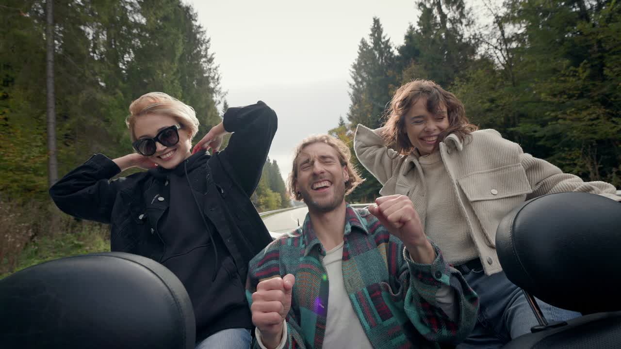 Two Girls And Young Man In Back Seat Of Car With An Open Roof. They Have Fun.