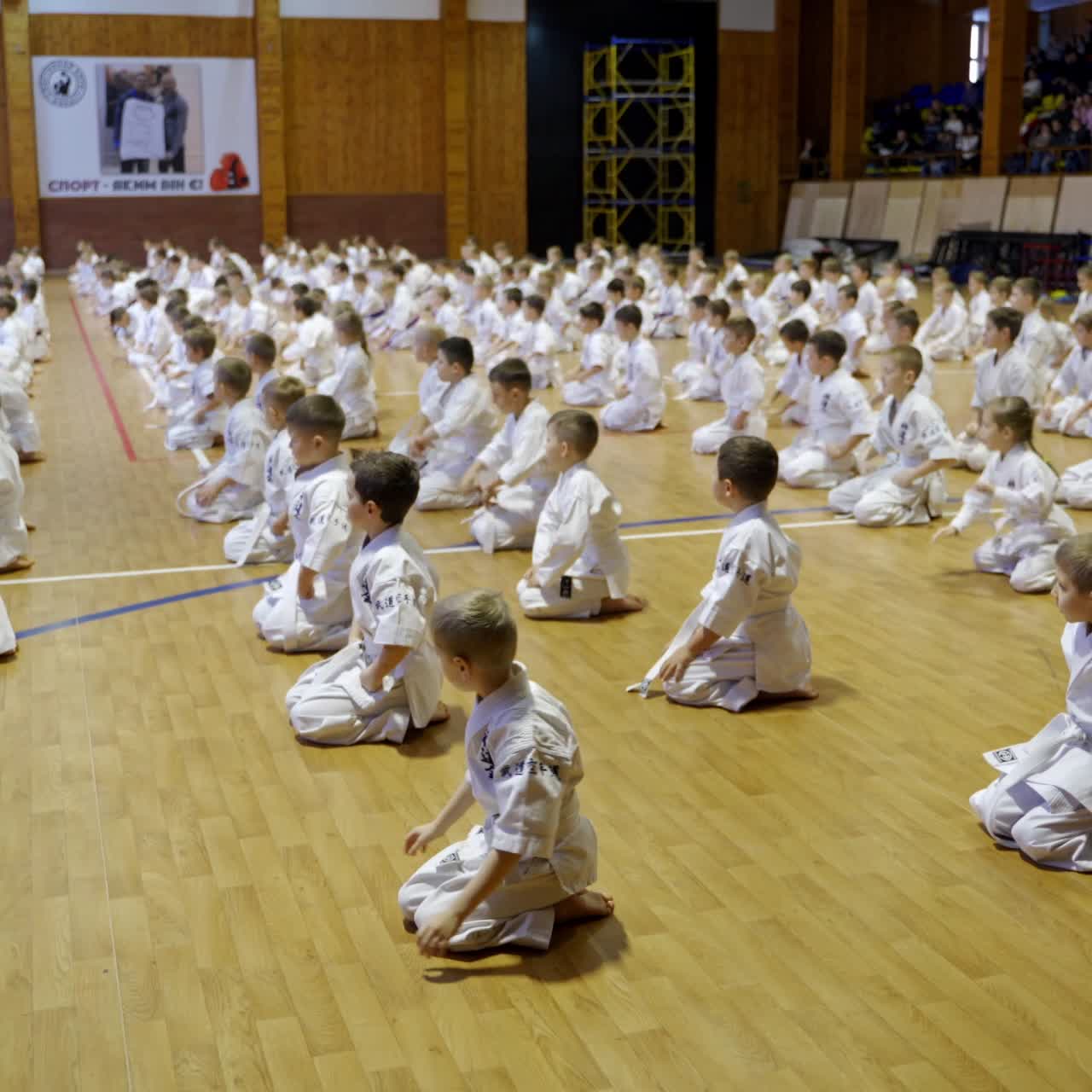 Large gym with numerous kids sitting on their knees on the floor. Children bend to greet the masters. Spectators sitting at the backdrop