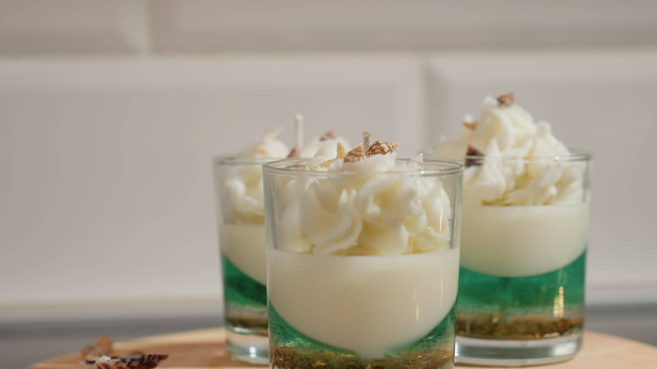 Top down view of beautifully crafted iced cake candle in glass cups, layered with green and white wax, topped with whipped textures and miniature shell decor, placed on wooden tray
