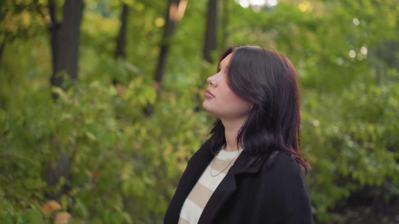 Medium shot of charming woman wearing long jacket walking slowly in forest, gazing around with serene thoughtful expression surrounded by lush greenery creating calm peaceful mood