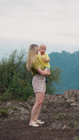 long haired woman prohibits little son to suck fingers standing on rocky hill top against distant mountain silhouettes at eco resort slow motion