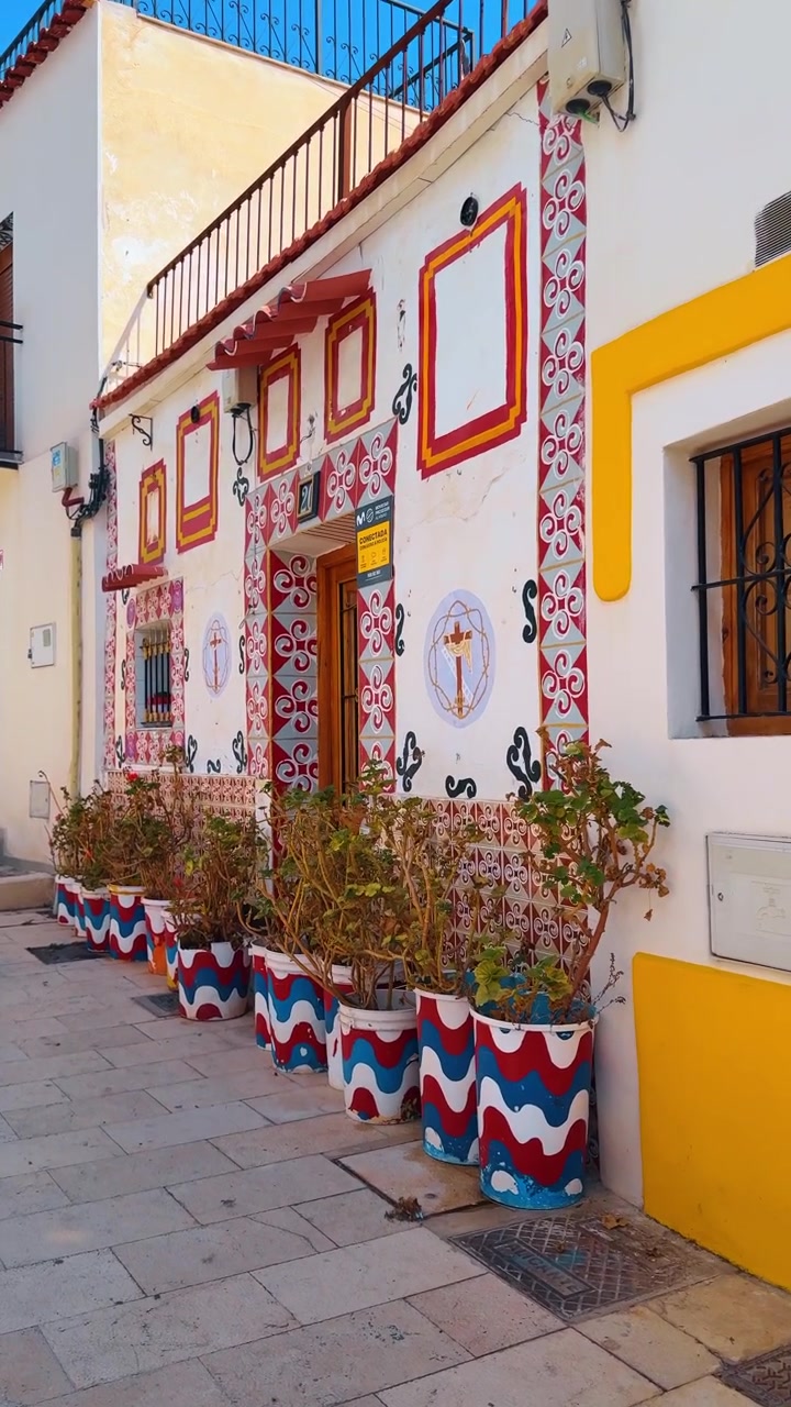 Santa Cruz Alicante'de beyaz evlerin olduğu dar sokak. İspanya'da beyaz binalar, sarı vurgular ve mavi, kırmızı, beyaz dalgalı desenli saksılarda bitkilerle geleneksel dar sokak.